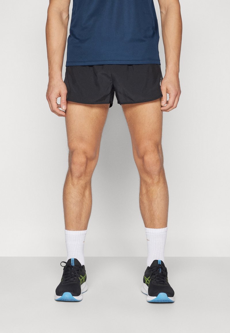Homme portant un short de course noir, un t-shirt bleu marine, des chaussettes blanches et des chaussures de course noires, debout devant un fond uni.