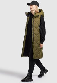 Olivgrüne gesteppte Weste mit hoch geschlossenem Kragen, Reißverschluss vorne, seitlichen Taschen, getragen über einem schwarzen Hoodie und schwarzen Hosen, kombiniert mit schwarzen Sneakers.