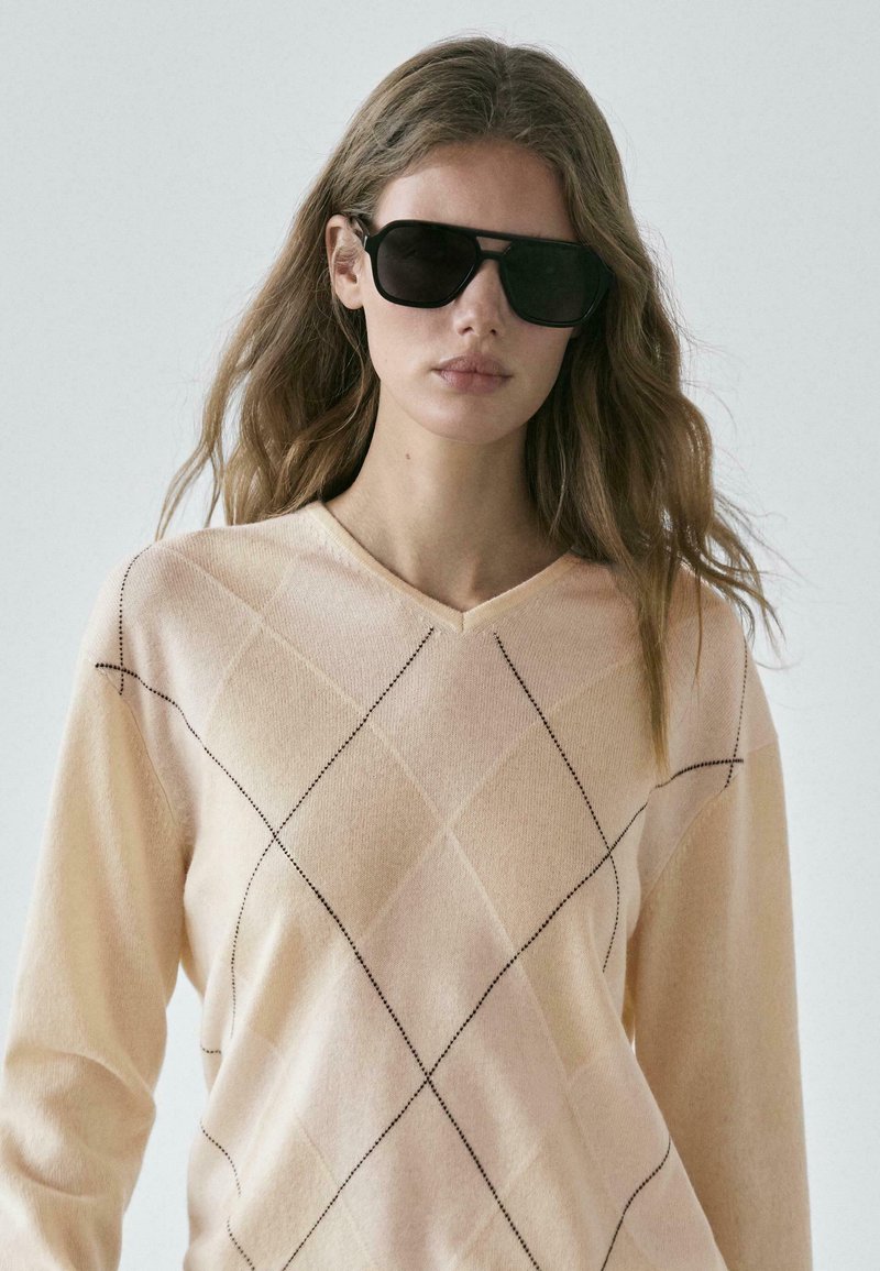 Pull rose clair à col en V avec un motif en losange en fil foncé. Le modèle porte de grandes lunettes de soleil noires et a de longs cheveux ondulés.