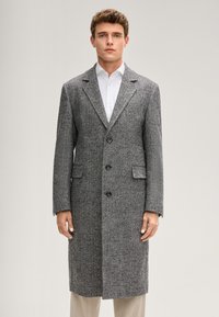 Cappotto grigio a spina di pesce con vestibilità sagomata, rever a lancia, tre bottoni e due tasche frontali. Indossato sopra una camicia bianca abbottonata.