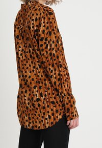 Camicia a fantasia leopardata marrone realizzata in tessuto testurizzato, con un design a bottoni, maniche lunghe e orlo curvo. Accentuata da macchie nere.