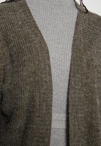 Olivově zelený svetr s texturovaným pleteným vzorem, otevřený vpředu a s širokými rukávy, vrstvený přes světle šedý žebrovaný rolák.