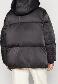 Giacca a vento nera con cappuccio, caratterizzata da un design trapuntato, polsini elastici e un fit ampio. Materiale liscio e lucido con una texture opaca.