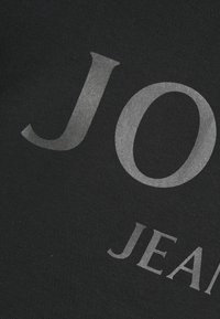 Tissu noir avec de grandes lettres argentées "JO" et de plus petites lettres argentées "JEAN" partiellement visibles, probablement une partie d'un logo ou d'un texte de marque.