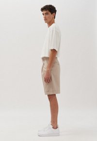 Shorts beige con pieghe, abbinati a una t-shirt bianca over. Il modello indossa sneakers bianche. Design semplice, colori chiari e texture in cotone.
