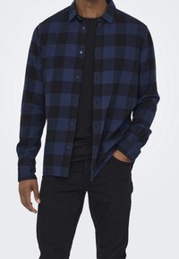 Camicia di flanella a quadri blu e neri con bottoni frontali, maniche lunghe e vestibilità comoda, abbinata a una t-shirt nera e jeans.