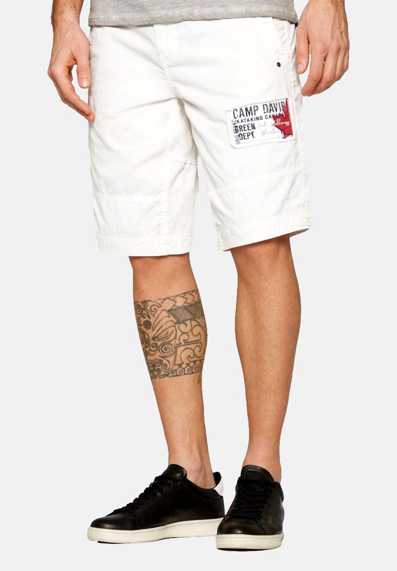 Șorturi cargo albe cu o patch-un care citește "CAMP DAVID" în roșu și negru. Purtați cu adidași negri; prezintă un tatuaj decorativ pe picior.
