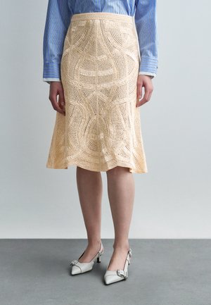 Mujer con falda beige de crochet hasta la rodilla, camisa de manga larga a rayas azules y zapatos blancos de tacón bajo con tira trasera.