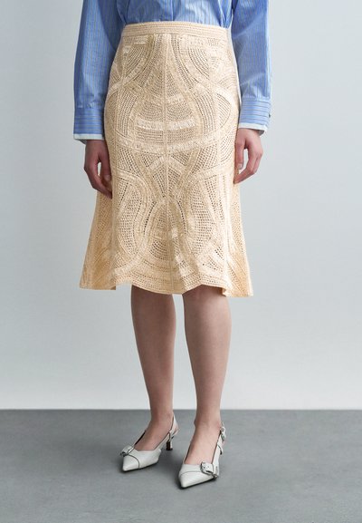 Femme portant une jupe beige en crochet longueur genou, une chemise à manches longues rayée bleue, et des chaussures blanches à talons bas et bride arrière.