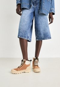 Bottines marron et crème avec une finition en daim texturé et une semelle en caoutchouc épaisse. La tige présente un laçage et des accents de doublure en fourrure.