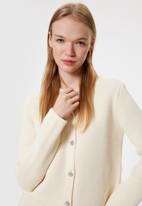 Crème cardigan met een gestructureerd breiselontwerp, voorzien van glanzende knopen en geribde boorden, weergegeven tegen een effen witte achtergrond.