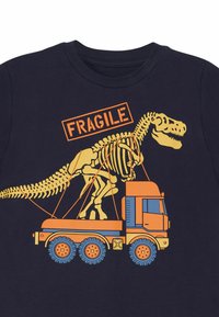 Marineblaues T-Shirt mit einem orangefarbenen Grafikdesign eines Dinosaurierskeletts auf einem Lastwagen, mit dem Wort "FRAGILE" darüber in fetter Schrift.