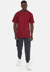 Mørkerød bomulds t-shirt med korte ærmer og et sort kursiv logo, parret med mørkegrå cargo-bukser og hvide sneakers.