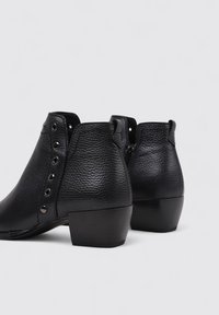 Botas de tobillo de cuero negro con un acabado texturizado, con un tacón bajo y bloque y ojetes metálicos circulares a lo largo del costado para los cordones.