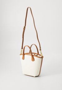 Kleine beige Canvas-Handtasche mit hellbraunen Ledergriffen, Besatz und verstellbarem langem Gurt mit goldfarbenen Beschlägen auf weißem Hintergrund.