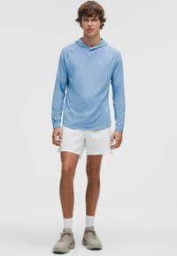 Lichtblauwe lange mouw hoodie met een subtiel gestructureerde stof, gecombineerd met witte shorts en lichtgrijze sneakers, staande tegen een effen achtergrond.