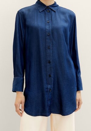 Camicia di jeans blu scuro con scollo a collo, chiusura frontale con bottoni e maniche lunghe. Presenta un orlo curvo e una texture liscia.