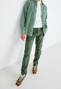 Chemise en velours côtelé vert sur un T-shirt blanc, associée à un pantalon vert et des baskets marron, avec des accents multicolores.