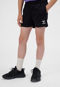Shorts atletici neri con vita elastica e tasche laterali. Presentano un logo bianco sulla gamba sinistra. Indossati con sneakers nere e calzini crema.