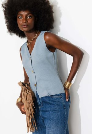 Femme aux cheveux afro naturels portant un haut bleu sans manches boutonné, un jean bleu, des bijoux en or, tenant un sac à franges beige sur fond blanc.