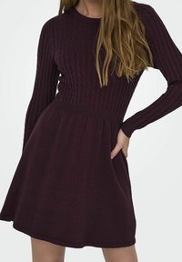 Femme portant une robe en tricot côtelé à manches longues, de couleur violet foncé, avec une taille cintrée et une jupe évasée, la main posée sur la hanche.