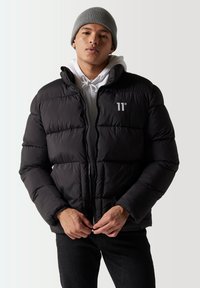 Chaqueta de plumas negra con diseño acolchado, cierre con cremallera y logo en el pecho. Llevada sobre una sudadera blanca, combinada con jeans oscuros y un gorro gris.