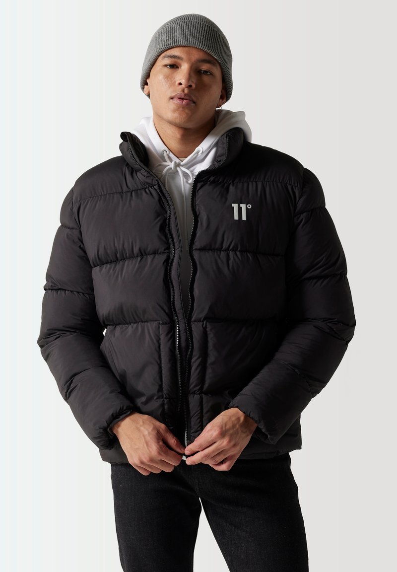 Chaqueta de plumas negra con diseño acolchado, cierre con cremallera y logo en el pecho. Llevada sobre una sudadera blanca, combinada con jeans oscuros y un gorro gris.