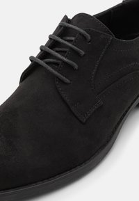 Chaussure en daim noir avec un bout arrondi, fermeture à lacets à six œillets et subtils accents de couture sur le dessus. Semelle plate en caoutchouc.