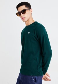 Donkergroene longsleeve t-shirt met een ronde halslijn en een klein wit logo op de borst, in combinatie met een donkere zonnebril.
