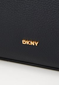 Melna ādas soma ar teksturētu virsmu un zelta "DKNY" logotipu, iegravētu priekšpusē. Kompakta forma ar tīrām līnijām.