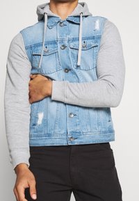 Chaqueta de mezclilla con un frente azul claro y mangas de sudadera gris. Cuenta con dos bolsillos en el pecho, botones en la parte delantera y un ligero desgaste en el dobladillo.
