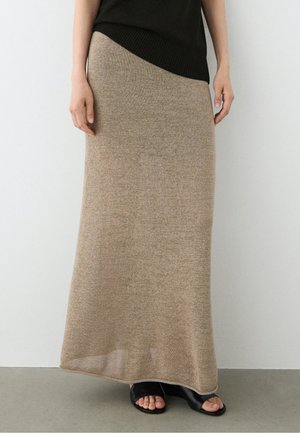 Femme portant une longue jupe en maille beige, un top noir et des sandales à talons noires à bout ouvert, debout contre un mur uni.