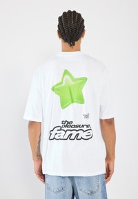Camiseta de algodón blanca con un gran gráfico de estrella verde en la parte posterior y texto negro. Corte relajado, mangas cortas y diseño de cuello redondo.