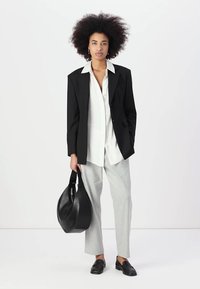 Blazer noir sur une chemise blanche à boutons, associé à un pantalon gris clair et des mocassins noirs. Tenant un sac à bandoulière noir, cheveux texturés.