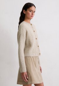 Beige Strickcardigan mit Rundhalsausschnitt und sechs braunen Knöpfen, kombiniert mit einem hellbeigen Rock, vor einem neutralen Hintergrund.