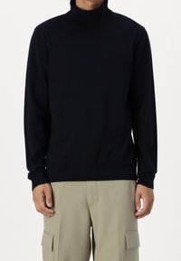 Man i svart långärmad polotröja och beige cargo-byxor, stående mot en enkel vit bakgrund.