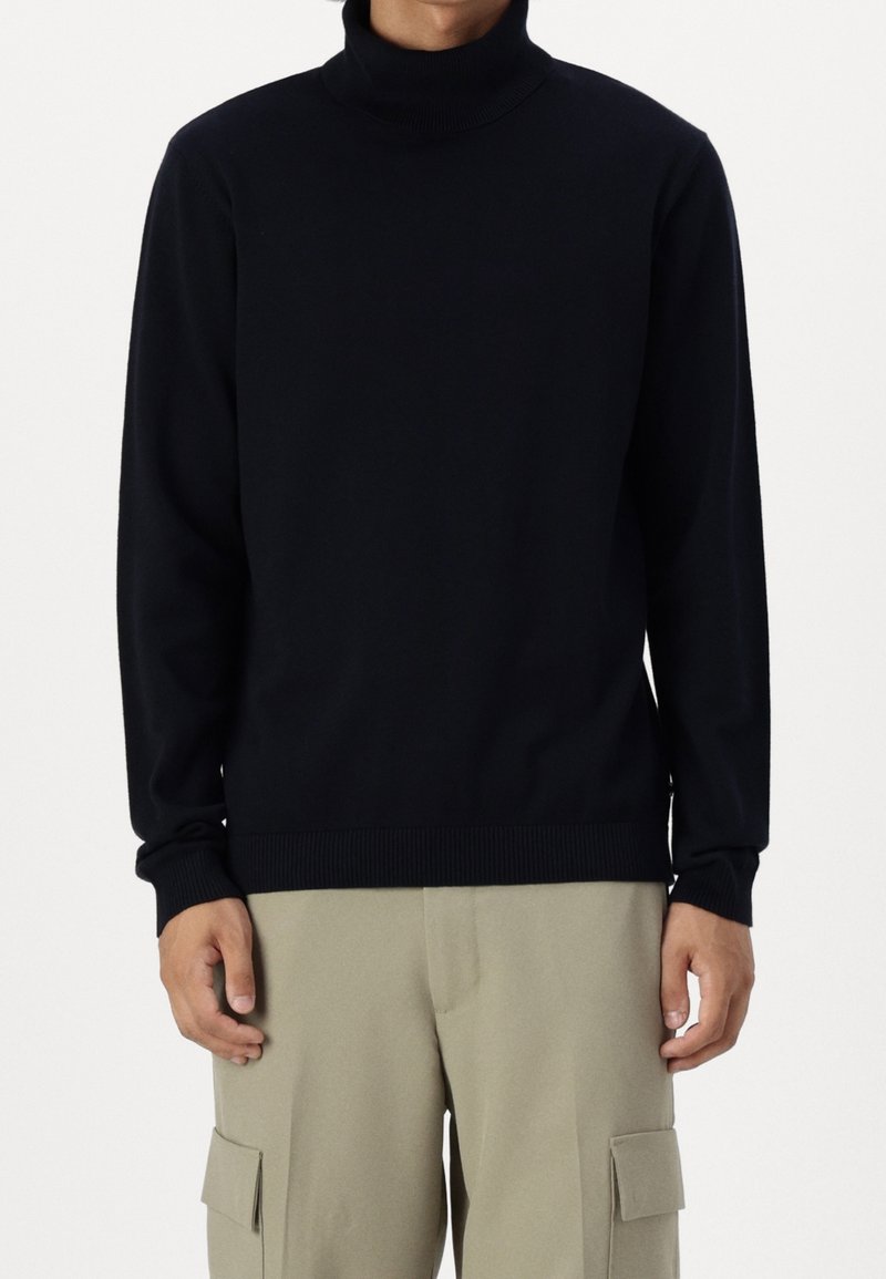 Man i svart långärmad polotröja och beige cargo-byxor, stående mot en enkel vit bakgrund.