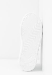 Semelle blanche de basket avec motif de grip texturé montrée verticalement contre un angle de mur et de sol blanc.