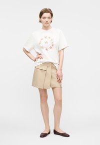 Ung kvinne iført hvit T-skjorte med blomsterprint i sirkel, beige plissert mini-skjørt og mørkebrune flate sko, står med hånden på hoften.