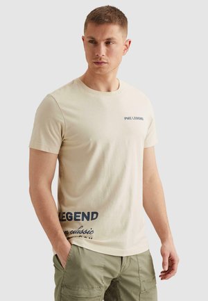 Mlad moški v bež majici PME Legend in olivno zelenih taktičnih hlačah, stoji z roko v žepu in gleda v stran.