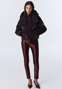 Doudoune bordeaux avec un col haut, fermeture éclair et poches avant, assortie à des leggings bordeaux brillants et des escarpins noirs pointus.