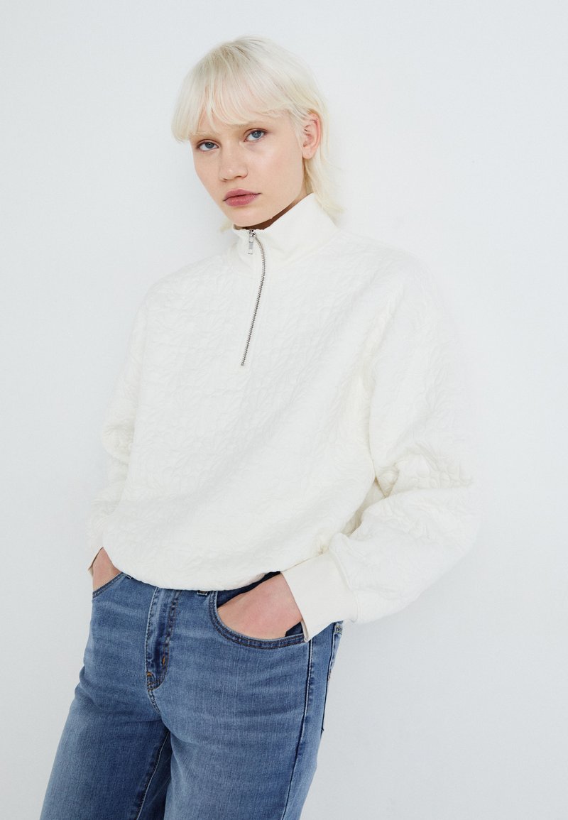 Weißer Reißverschluss-Sweatshirt mit strukturiertem Design, hohem Kragen und langen Ärmeln, kombiniert mit blauen Jeans. Schlichte und moderne Ästhetik.
