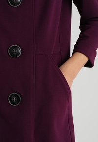 Burgundy kappa i en strukturerad tyg, med stora svarta knappar, en sidficka och trekvartslånga ärmar.