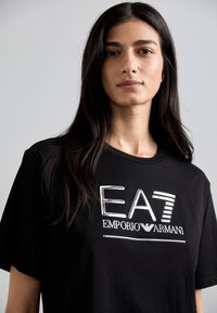 Melna kokvilnas T-krekls ar lielu sudraba logotipu, kas lasāms "EA7 EMPORIO ARMANI". Klasiskais griezums ar apaļu kakla izgriezumu un īsām piedurknēm.