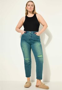 Černý tílko bez rukávů spárované s vysokými modrými džíny s poškozenými detaily a světle zelenými akcenty, doplněné béžovými slip-on botami.
