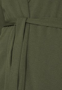 Vestido de envoltura verde oliva hecho de tejido texturizado, con un cinturón de lazo y un acabado suave. La vista en primer plano resalta el material y el color.
