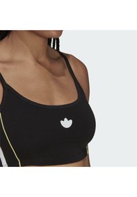 Kvinna iklädd en svart sport-bh med tunna remmar och en vit logotyp centrerad på bröstet mot en enfärgad ljus bakgrund.