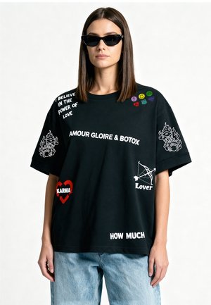 Femme portant un T-shirt noir oversize avec des patchs de texte blancs et rouges, un jean bleu clair et des lunettes de soleil noires œil-de-chat sur fond blanc.