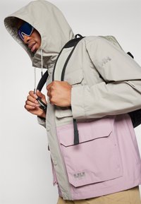 Hellgraue Jacke mit lila Akzenten, ausgestattet mit einer Kapuze, Taschen und einer strukturierten Oberfläche. Zeigt das Logo auf der Vordertasche.