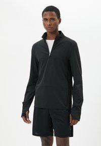 Pullover atletico nero con zip a metà in tessuto leggero, maniche lunghe e aperture per il pollice, abbinato a pantaloni corti neri, in piedi contro uno sfondo bianco.